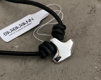 May include: A silver-colored pendant in the shape of a hammer is strung on a black cord. The pendant is the focal point, with a tag that reads "BARBARIANN history inspired jewelry" in the background.
