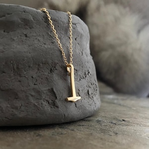 Può includere: Una collana con pendente dorato con un piccolo design a martello. Il pendente è appeso a una delicata catena d'oro. La collana è esposta su una pietra grigio scuro e strutturata. Lo sfondo è sfocato, con un accenno di grigio chiaro.