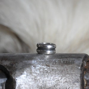 May include: A silver ring with two bands, one slightly larger than the other, resting on a metal surface.