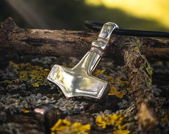 May include: A silver Mjolnir pendant, a Norse symbol of strength and protection, hangs on a black leather cord. The pendant is resting on a piece of wood with yellow lichen.