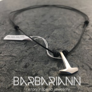 May include: A silver Thor's hammer pendant on a black leather cord necklace. The pendant is a simple, geometric design with a smooth, polished finish. The necklace is tied with a knot at the back.