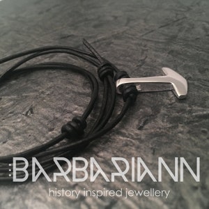 May include: A black leather bracelet with a silver anchor charm. The bracelet is tied with a knot and has a braided leather design. The text "BARBARIANN history inspired jewellery" is visible at the bottom of the image.
