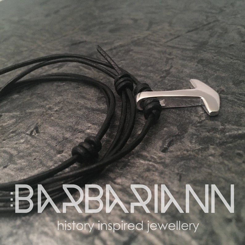 May include: A black leather bracelet with a silver anchor charm. The bracelet is tied with a knot and has a braided leather design. The text "BARBARIANN history inspired jewellery" is visible at the bottom of the image.