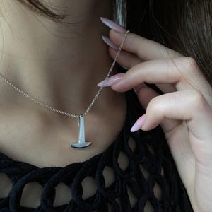 Könnte beinhalten: Ein silberfarbener Anhänger in Form eines stilisierten Hammers, der an einer zarten, passenden silberfarbenen Kette hängt. Der Anhänger hat eine glatte, polierte Metalloberfläche mit einem klaren, geometrischen Design, das vertikal an der Kette hängt.