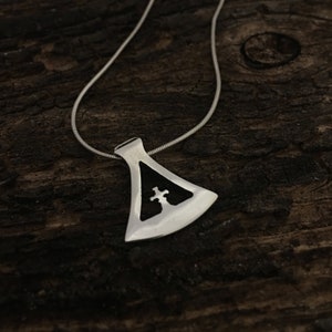 Puede incluir: Un collar de plata con un colgante en forma de hacha con una cruz en su interior. El colgante está sobre una superficie de madera.