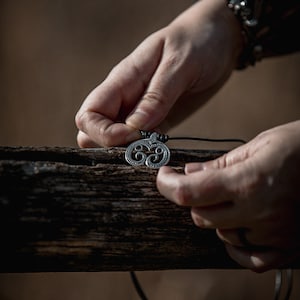 Puede incluir: Un colgante de plata con un diseño de nudo celta cuelga de un cordón negro. El colgante está siendo sostenido por la mano de una persona.