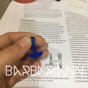 May include: A close-up of a blue Thor's hammer pendant. The pendant is made of resin and is a replica of a Viking age artifact. The pendant is being held in a hand. The image also shows a line drawing of a similar Thor's hammer pendant.
