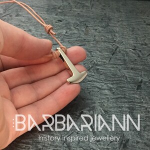 Könnte beinhalten: Ein silberfarbener Anhänger in Form eines Hammers, der an einem hellbraunen Lederband hängt. Der Anhänger befindet sich auf einer Hand vor einem grauen Hintergrund. Der Text "BARBARIANN history inspired jewellery" ist am unteren Rand des Bildes sichtbar.