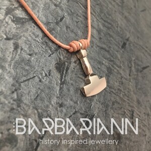 Puede incluir: Un colgante plateado con forma de martillo, suspendido de un cordón marrón claro. El colgante está detallado con un mango y una cabeza de martillo. El cordón está anudado en la parte superior. El texto "BARBARIANN history inspired jewellery" está en la parte inferior.