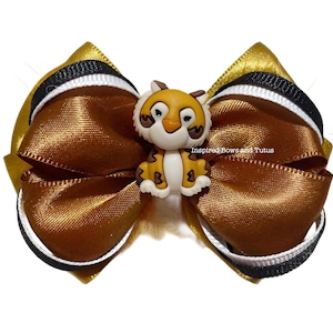 May include: A handmade hair bow featuring a gold and brown satin ribbon design with black and white trim. A small, cartoon tiger figurine is centered on the bow. The bow is a decorative hair accessory.
