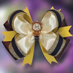May include: A handmade hair bow featuring a cream-colored satin center, accented with yellow, brown, and navy blue ribbons. The bow is embellished with gold glitter and a small clock-shaped charm. The text "Inspired Bows and Tutus" is visible.