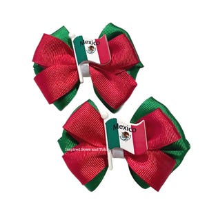 May include: Two hair bows with red and green ribbon, each featuring a small Mexican flag. The bows have a layered design with a central flag element. The bows are likely for festive occasions.