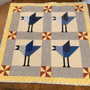 May include: A quilted wall hanging with a yellow border and a pattern of blue birds with black legs and beaks on a beige background.