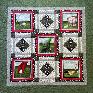 May include: A golf-themed quilt with nine squares. Each square features golf imagery, including golf balls, clubs, and a golf bag. The squares are framed in red and black, set against a light gray background with a green border. The quilt is approximately 40 inches square.