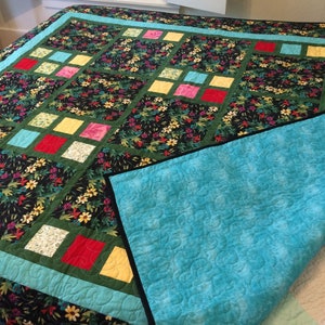 May include: A handmade quilt with a floral pattern in black, green, and yellow. The quilt features colorful square accents in red, pink, yellow, and blue. The underside is a solid turquoise color. The quilt is displayed on a bed.