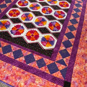 May include: A quilt with a patchwork design featuring a repeating pattern of colorful flowers on a black background. The quilt is bordered with a purple and white diamond pattern.