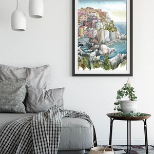 K&ouml;nnte beinhalten: Aquarellmalerei eines K&uuml;stendorfes mit farbenfrohen Geb&auml;uden auf einer Klippe, schwarz gerahmt. Das Kunstwerk wird in einem Wohnzimmer mit einem grauen Sofa, Kissen und einer gemusterten Decke ausgestellt.