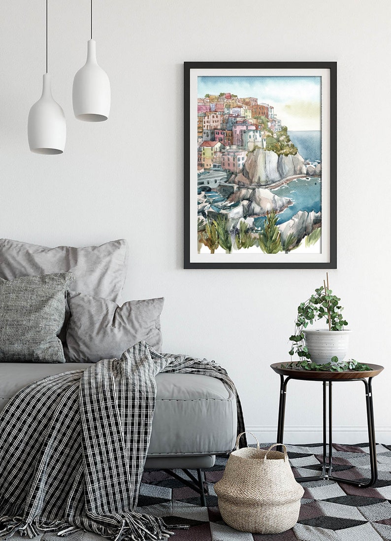 K&ouml;nnte beinhalten: Aquarellmalerei eines K&uuml;stendorfes mit farbenfrohen Geb&auml;uden auf einer Klippe, schwarz gerahmt. Das Kunstwerk wird in einem Wohnzimmer mit einem grauen Sofa, Kissen und einer gemusterten Decke ausgestellt.