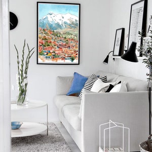 Pu&ograve; includere: Un divano bianco con cuscini blu e bianco e nero in un soggiorno con un dipinto ad acquerello incorniciato di una citt&agrave; con montagne innevate sullo sfondo. C'&egrave; un tavolino da caff&egrave; bianco e un porta riviste bianco con riviste davanti al divano.