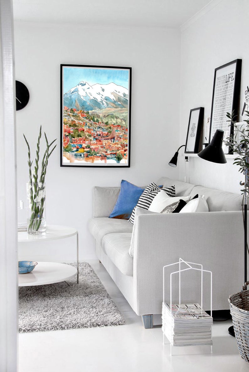 Pu&ograve; includere: Un divano bianco con cuscini blu e bianco e nero in un soggiorno con un dipinto ad acquerello incorniciato di una citt&agrave; con montagne innevate sullo sfondo. C'&egrave; un tavolino da caff&egrave; bianco e un porta riviste bianco con riviste davanti al divano.