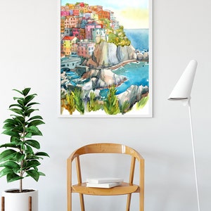K&ouml;nnte beinhalten: Aquarellmalerei eines farbenfrohen K&uuml;stendorfes mit Geb&auml;uden auf einer Klippe, mit Blick auf das blaue Meer. Das Kunstwerk ist wei&szlig; gerahmt und an einer wei&szlig;en Wand &uuml;ber einem Holzstuhl und einer Topfpflanze ausgestellt.