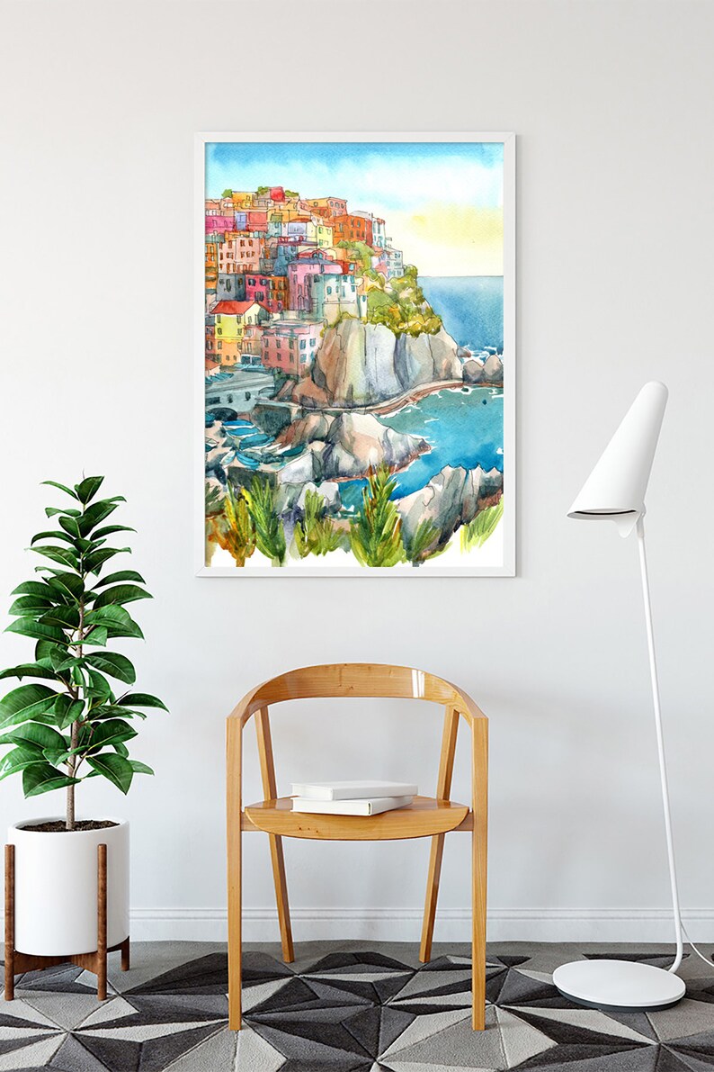 K&ouml;nnte beinhalten: Aquarellmalerei eines farbenfrohen K&uuml;stendorfes mit Geb&auml;uden auf einer Klippe, mit Blick auf das blaue Meer. Das Kunstwerk ist wei&szlig; gerahmt und an einer wei&szlig;en Wand &uuml;ber einem Holzstuhl und einer Topfpflanze ausgestellt.