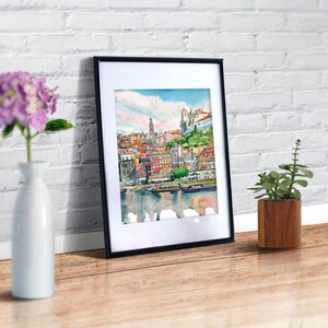 K&ouml;nnte beinhalten: Ein gerahmtes Aquarellbild einer Stadtlandschaft mit Geb&auml;uden, einem Fluss und Booten. Das Bild ist in einem schwarzen Rahmen und h&auml;ngt an einer wei&szlig;en Backsteinwand.