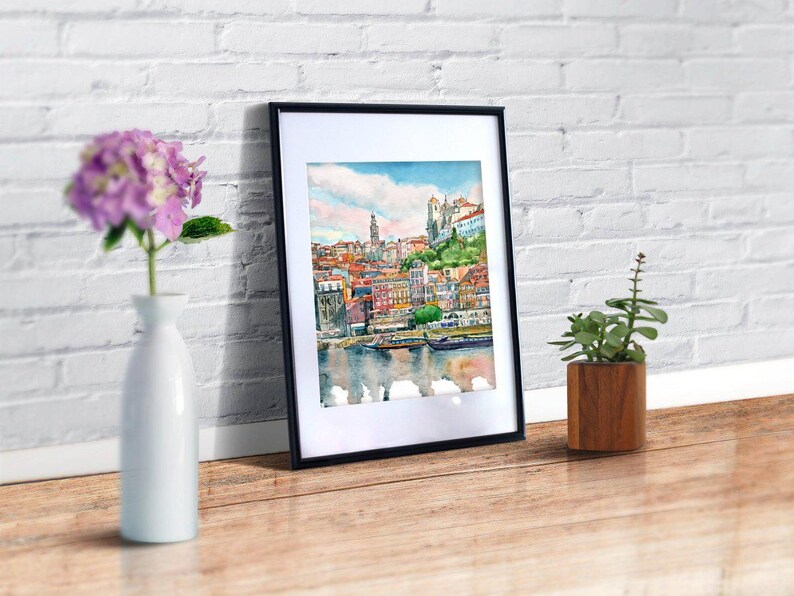 K&ouml;nnte beinhalten: Ein gerahmtes Aquarellbild einer Stadtlandschaft mit Geb&auml;uden, einem Fluss und Booten. Das Bild ist in einem schwarzen Rahmen und h&auml;ngt an einer wei&szlig;en Backsteinwand.