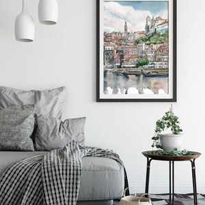 K&ouml;nnte beinhalten: Ein Aquarellbild einer Stadtlandschaft mit Geb&auml;uden, einem Fluss und Booten. Das Bild ist in einem schwarzen Rahmen gerahmt und h&auml;ngt an einer wei&szlig;en Wand.
