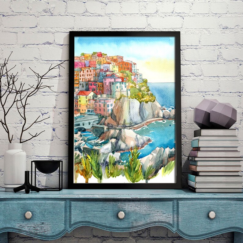 K&ouml;nnte beinhalten: Aquarellmalerei eines K&uuml;stendorfes mit farbenfrohen Geb&auml;uden auf einer Klippe, schwarz gerahmt. Das Kunstwerk zeigt eine lebendige Szene mit blauem Wasser, gr&uuml;nem Laub und klarem Himmel. Das Gem&auml;lde wird auf einem blauen Tisch ausgestellt.