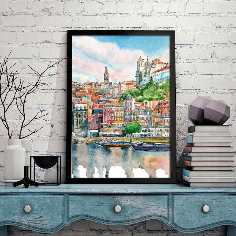 K&ouml;nnte beinhalten: Aquarellgem&auml;lde einer Stadtlandschaft mit bunten Geb&auml;uden, einer Kirche und Booten auf einem Fluss. Das Gem&auml;lde ist in Schwarz gerahmt und auf einem blauen Regal ausgestellt.