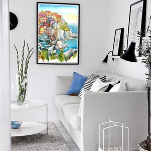 K&ouml;nnte beinhalten: Wohnzimmer-Interieur mit einem gerahmten Aquarellgem&auml;lde eines K&uuml;stendorfes. Ein graues Sofa mit blauen und gemusterten Kissen steht neben einem wei&szlig;en Couchtisch und einem flauschigen Teppich. Eine schwarze Uhr und Drucke schm&uuml;cken die wei&szlig;en W&auml;nde.