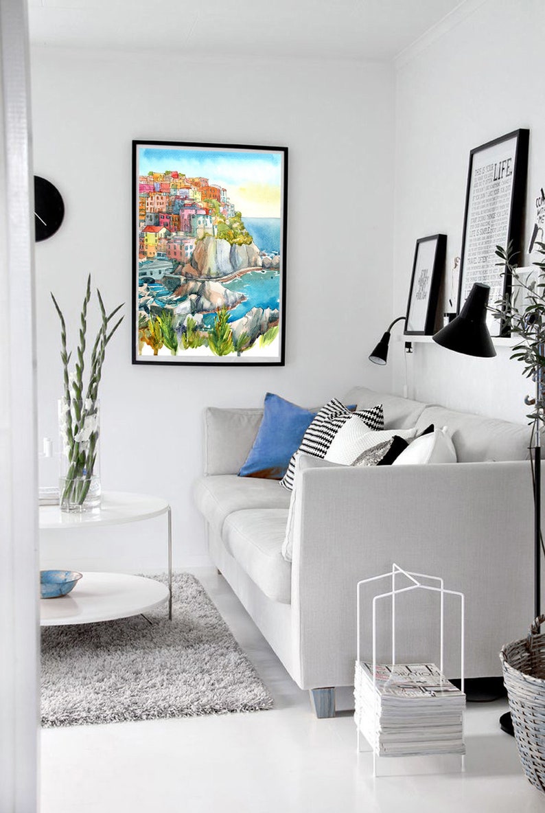 K&ouml;nnte beinhalten: Wohnzimmer-Interieur mit einem gerahmten Aquarellgem&auml;lde eines K&uuml;stendorfes. Ein graues Sofa mit blauen und gemusterten Kissen steht neben einem wei&szlig;en Couchtisch und einem flauschigen Teppich. Eine schwarze Uhr und Drucke schm&uuml;cken die wei&szlig;en W&auml;nde.