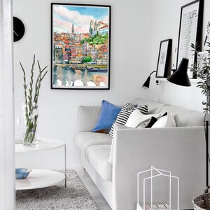 K&ouml;nnte beinhalten: Ein Aquarellbild einer Stadtlandschaft mit bunten Geb&auml;uden und einem Fluss im Vordergrund. Das Bild ist gerahmt und h&auml;ngt an einer wei&szlig;en Wand in einem Wohnzimmer. Eine wei&szlig;e Couch mit blauen und wei&szlig;en Kissen steht im Vordergrund.