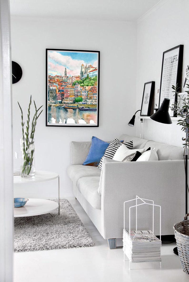 K&ouml;nnte beinhalten: Ein Aquarellbild einer Stadtlandschaft mit bunten Geb&auml;uden und einem Fluss im Vordergrund. Das Bild ist gerahmt und h&auml;ngt an einer wei&szlig;en Wand in einem Wohnzimmer. Eine wei&szlig;e Couch mit blauen und wei&szlig;en Kissen steht im Vordergrund.