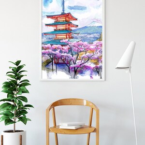May include: Watercolor print of a Japanese pagoda with cherry blossoms in a white frame. The artwork features a red and teal pagoda, pink blossoms, and a mountain in the background. The print is displayed above a wooden chair and a potted plant.