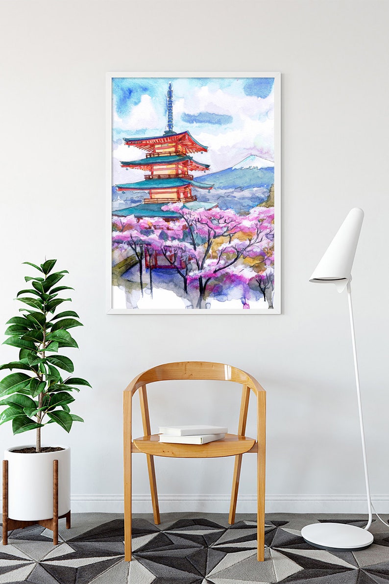 May include: Watercolor print of a Japanese pagoda with cherry blossoms in a white frame. The artwork features a red and teal pagoda, pink blossoms, and a mountain in the background. The print is displayed above a wooden chair and a potted plant.