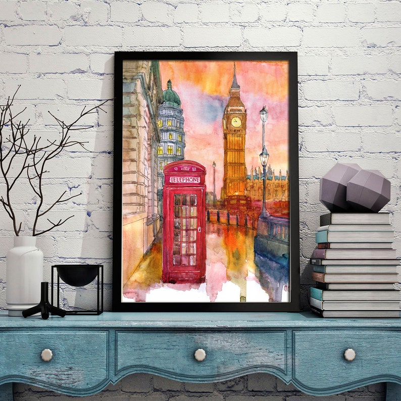 May include: Watercolor painting of Big Ben and a red telephone booth in London, England. The painting has a sunset sky with orange, pink, and purple hues. The painting is framed in black.