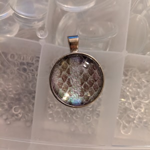 May include: A circular silver-toned pendant featuring a domed glass cabochon with an iridescent, scale-like pattern in shades of purple, green, and silver. The pendant has a simple metal bail at the top for attachment to a chain or cord.
