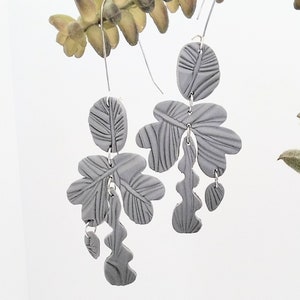 May include: A pair of gray dangle earrings with a leaf design. The earrings are made of polymer clay and have a textured surface.