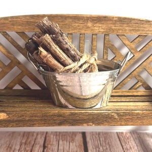 May include: A miniature wooden bench with a lattice back design and a weathered brown finish. Resting on the seat is a small, silver-colored metal bucket filled with a bundle of thin, dark brown twigs tied together with a piece of natural twine.