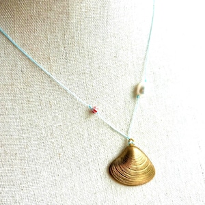 May include: A gold-colored seashell pendant necklace with a light blue cord. The necklace features a small seashell bead and two red and white beads. The pendant is a detailed, textured seashell.