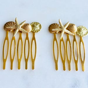 May include: Two gold-colored hair combs, each adorned with a starfish, seashell, and sand dollar design. The combs have four tines each. The decorative elements are in a warm, metallic tone, evoking a beach or nautical theme.