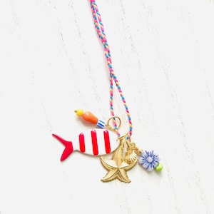 May include: A necklace featuring a braided cord in pink, blue, and orange, adorned with a white and red striped fish charm, a gold-toned starfish charm, a small seashell charm, and several colorful ceramic beads in blue, orange, and yellow.