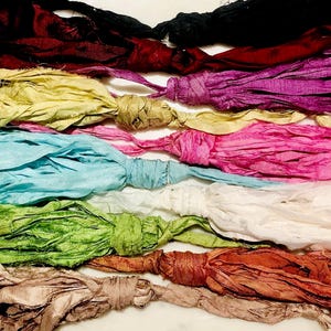 Op de afbeelding: Een verzameling kleurrijke zijden kwasten in verschillende tinten, waaronder zwart, bordeauxrood, paars, geel, roze, turquoise, wit, groen, oranje en beige. Elke kwast is bovenaan geknoopt.