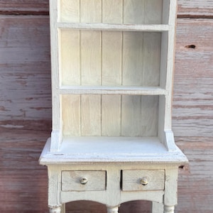 May include: A miniature wooden hutch painted in a distressed off-white finish. The piece features an open top section with two shelves, a central counter area, two small drawers with silver-toned knobs, and a lower shelf supported by four legs.