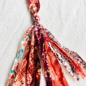 Puede incluir: Un borla hecha a mano con una parte superior anudada y múltiples tiras de tela. Las tiras muestran patrones florales y paisley en tonos de rojo, rosa, naranja y azul. La borla es un elemento decorativo.