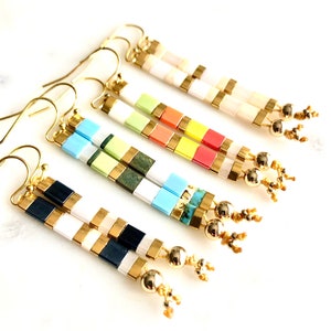 Puede incluir: Cinco pares de pendientes geométricos coloridos con detalles dorados. Cada par presenta una combinación de colores diferente de azulejos cuadrados, incluyendo blanco, negro, azul, verde, amarillo, naranja y rojo.