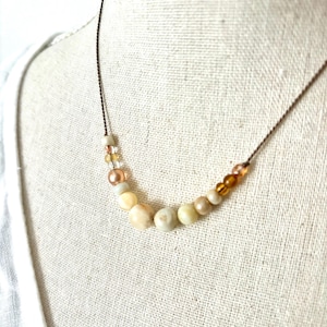 May include: A delicate necklace featuring a variety of beads in shades of cream, peach, and amber. The beads are strung on a thin, brown cord. The necklace is displayed on a white background, suggesting a minimalist aesthetic.