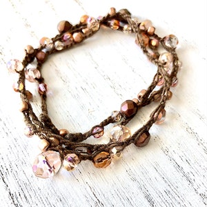 May include: A handcrafted bracelet with a brown cord, adorned with iridescent beads in pink, peach, and brown. The beads vary in size and are strung along the cord, creating a layered design. The bracelet is presented on a white, textured surface.